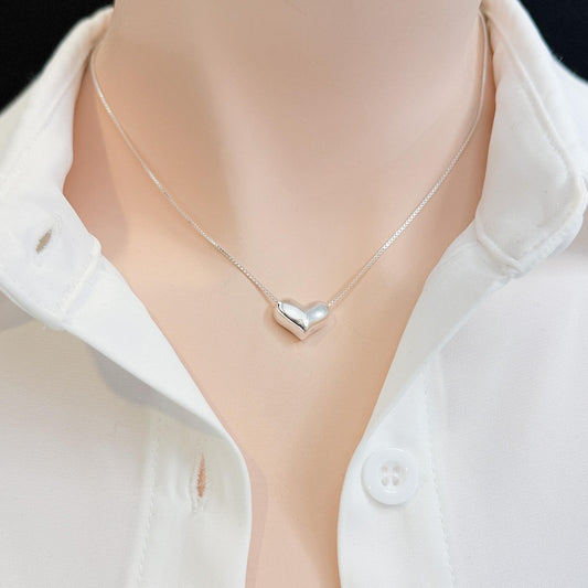 A silver heart-shaped pendant necklace is displayed on a person's neck, wearing a white collared shirt.