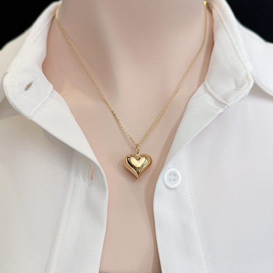 A gold heart-shaped pendant necklace is worn around a person's neck, with a white collared shirt visible underneath.