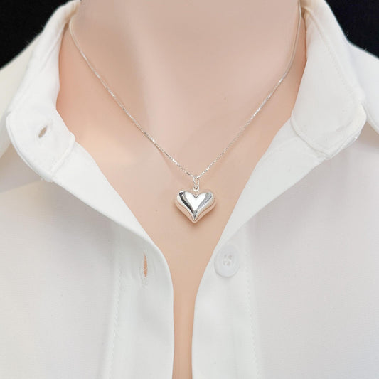 A silver heart-shaped pendant necklace is worn around a person's neck, with a white collared shirt visible underneath.