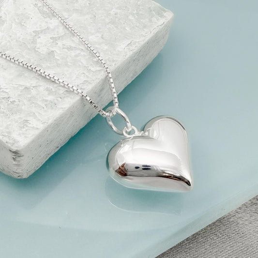 A silver heart-shaped pendant necklace is displayed on a white stone surface.