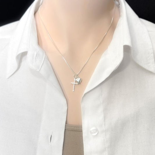 A silver necklace with a cross pendant and a small heart charm is worn around a person's neck.