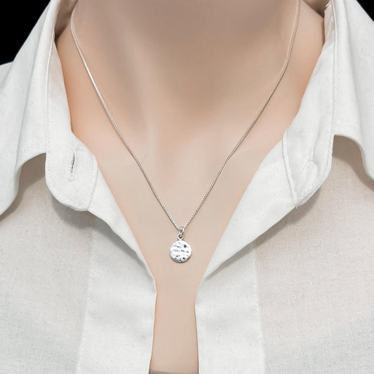 A silver necklace with a round pendant is worn around a person's neck, who is wearing a white collared shirt.