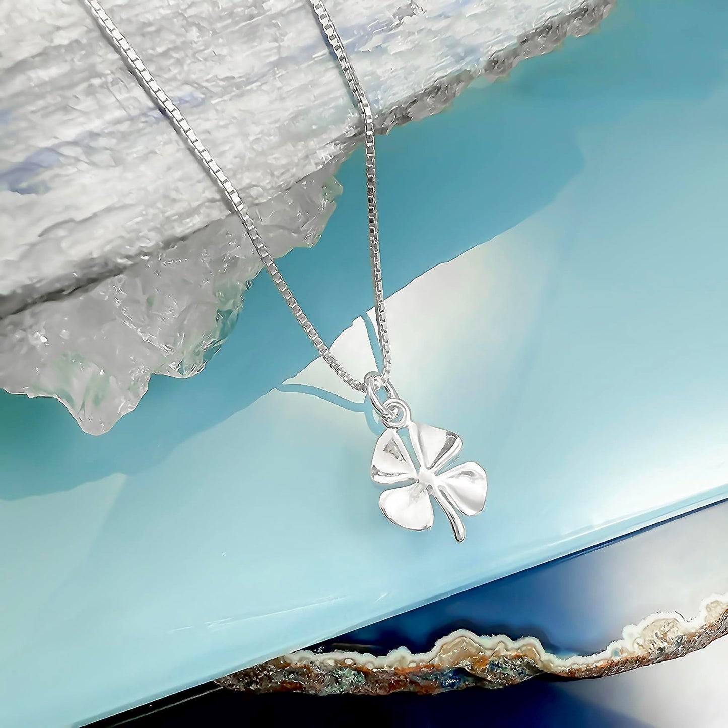 A silver-colored necklace with a small silver clover pendant is displayed against a blue background.