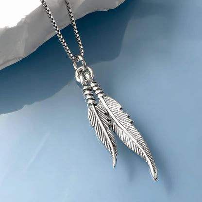 A silver-toned pendant necklace with two feather-like charms hanging from a chain against a blue background.