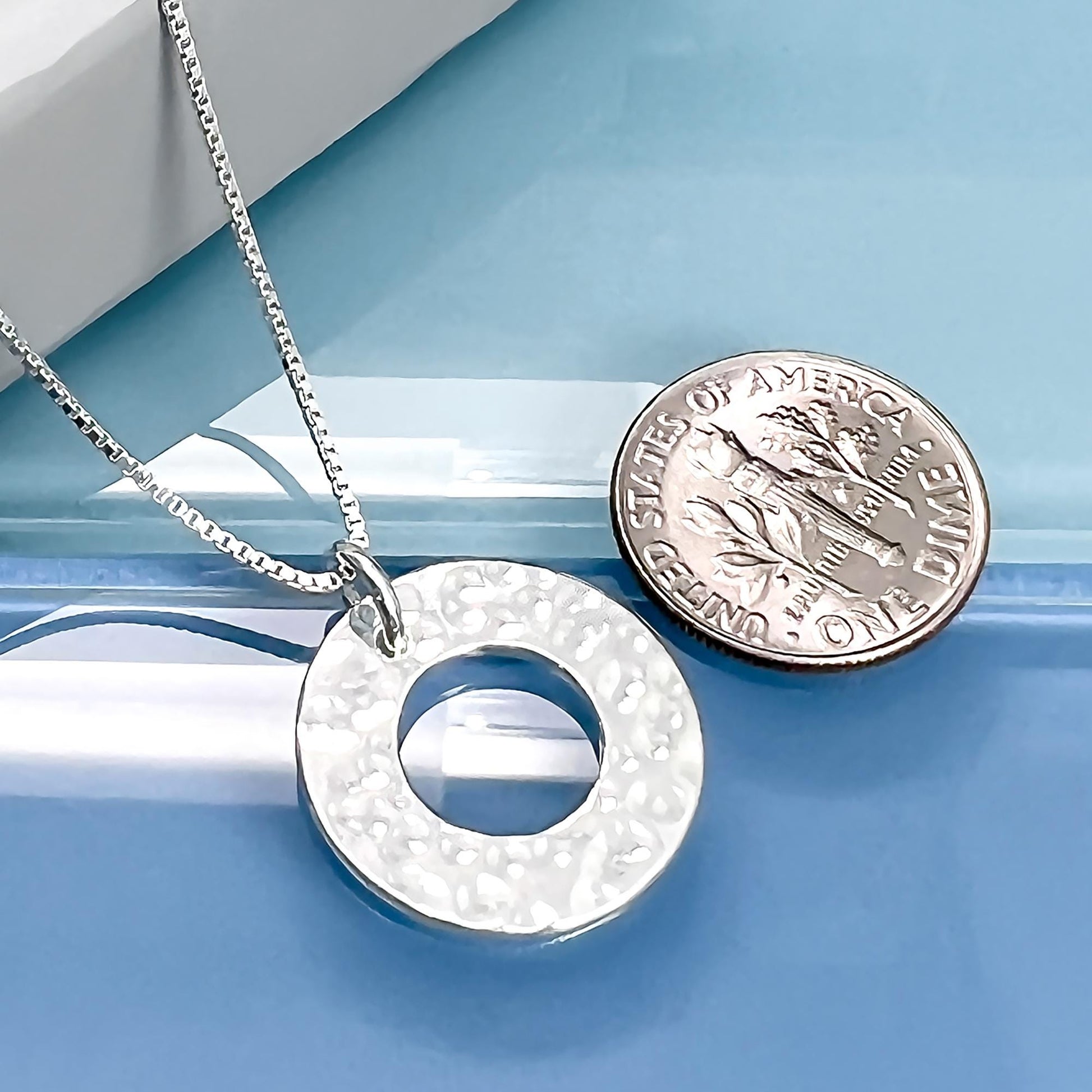 A silver necklace with a circular pendant is placed next to a United States one dollar coin on a blue surface.