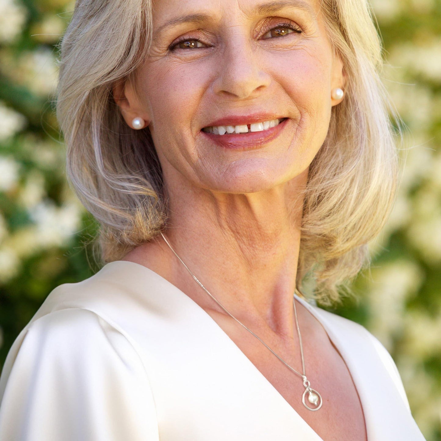 A smiling woman with blonde hair wearing a white blouse and a necklace with a pendant.