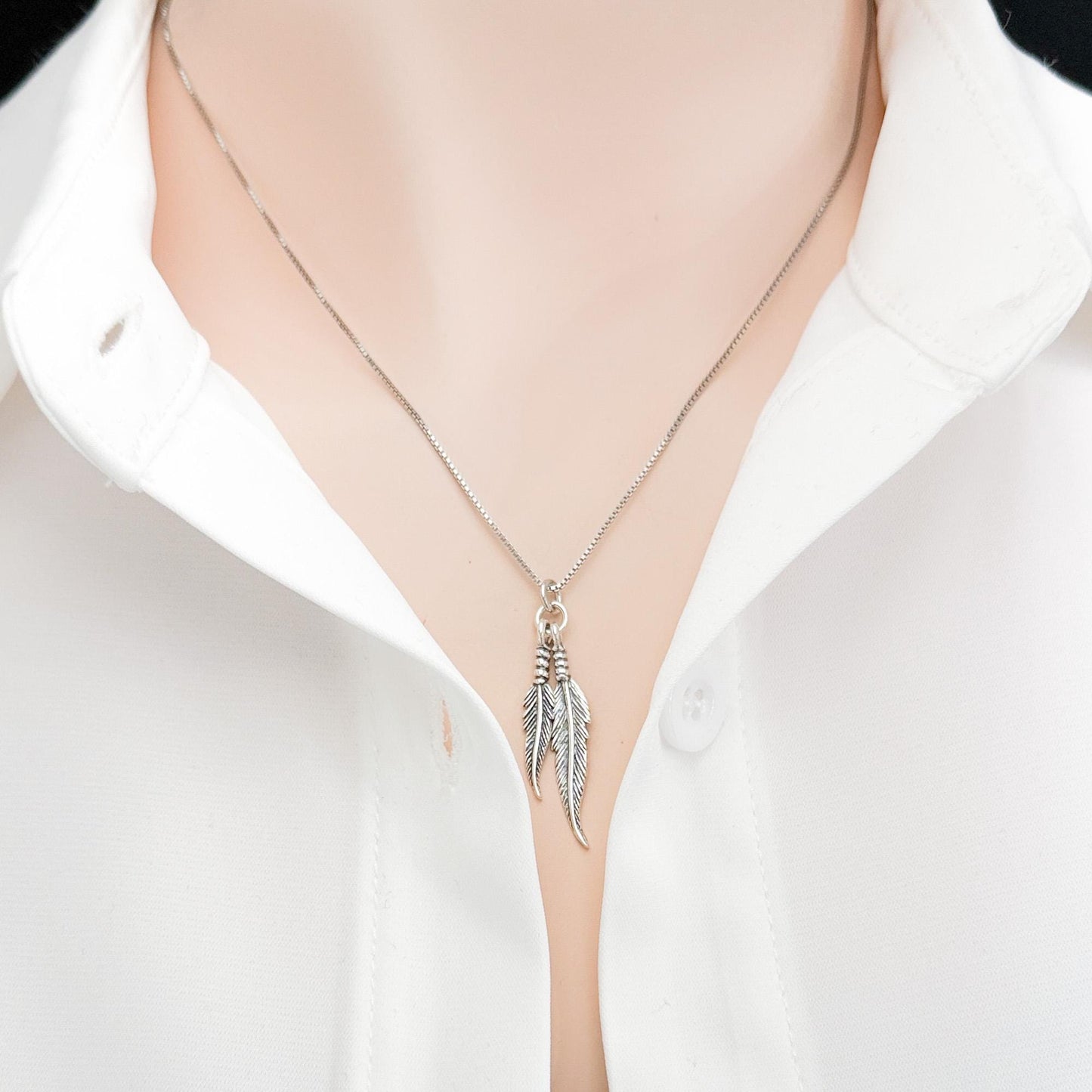A silver necklace with a pendant featuring feathers is shown against a white shirt collar.