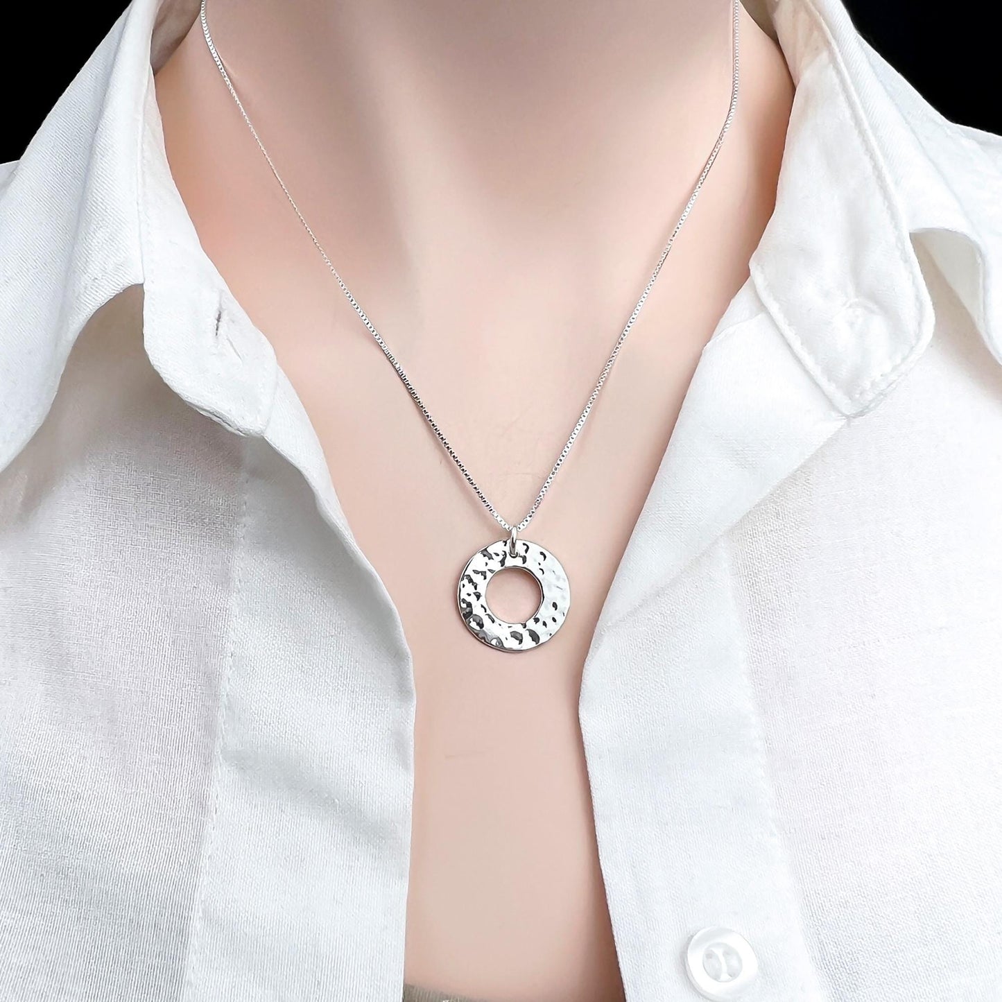 A silver necklace with a circular pendant is worn around a person's neck, who is wearing a white collared shirt.