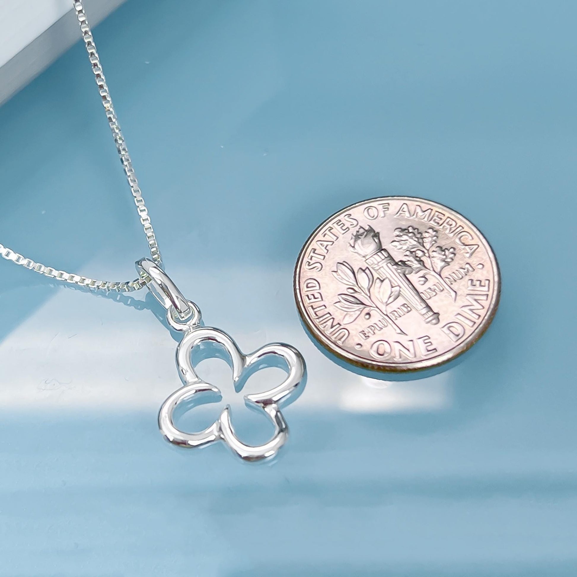 A silver flower pendant necklace and a United States one dollar coin are placed on a blue surface.