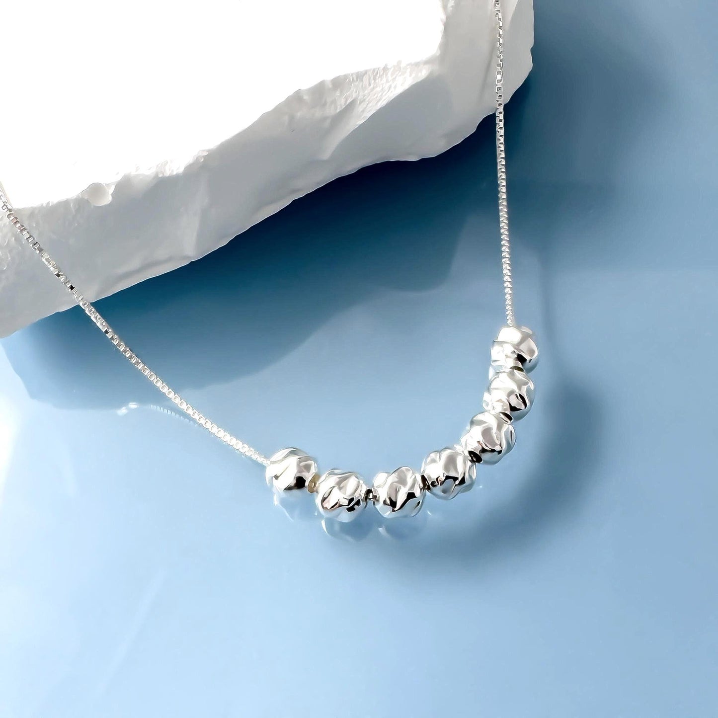 A silver necklace with a row of small, round, shiny beads is displayed against a blue background.