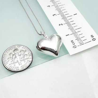 a silver heart-shaped locket necklace and a United States one dollar coin, with a ruler placed next to them.