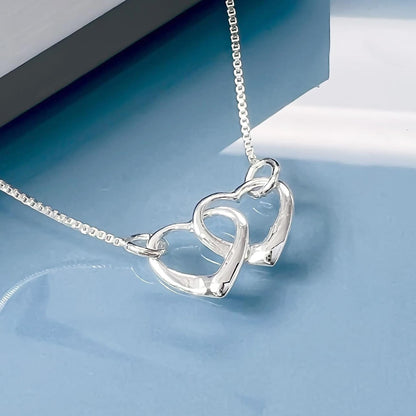 A silver necklace with two heart-shaped pendants is displayed against a blue background.