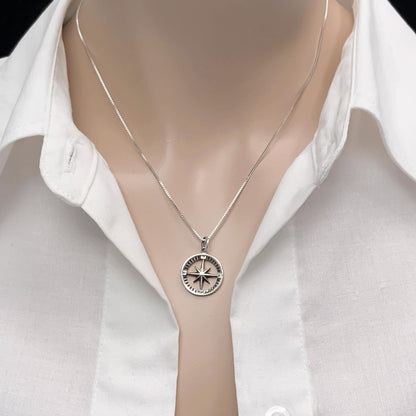 A silver necklace with a compass pendant is worn around a person's neck, who is wearing a white collared shirt.