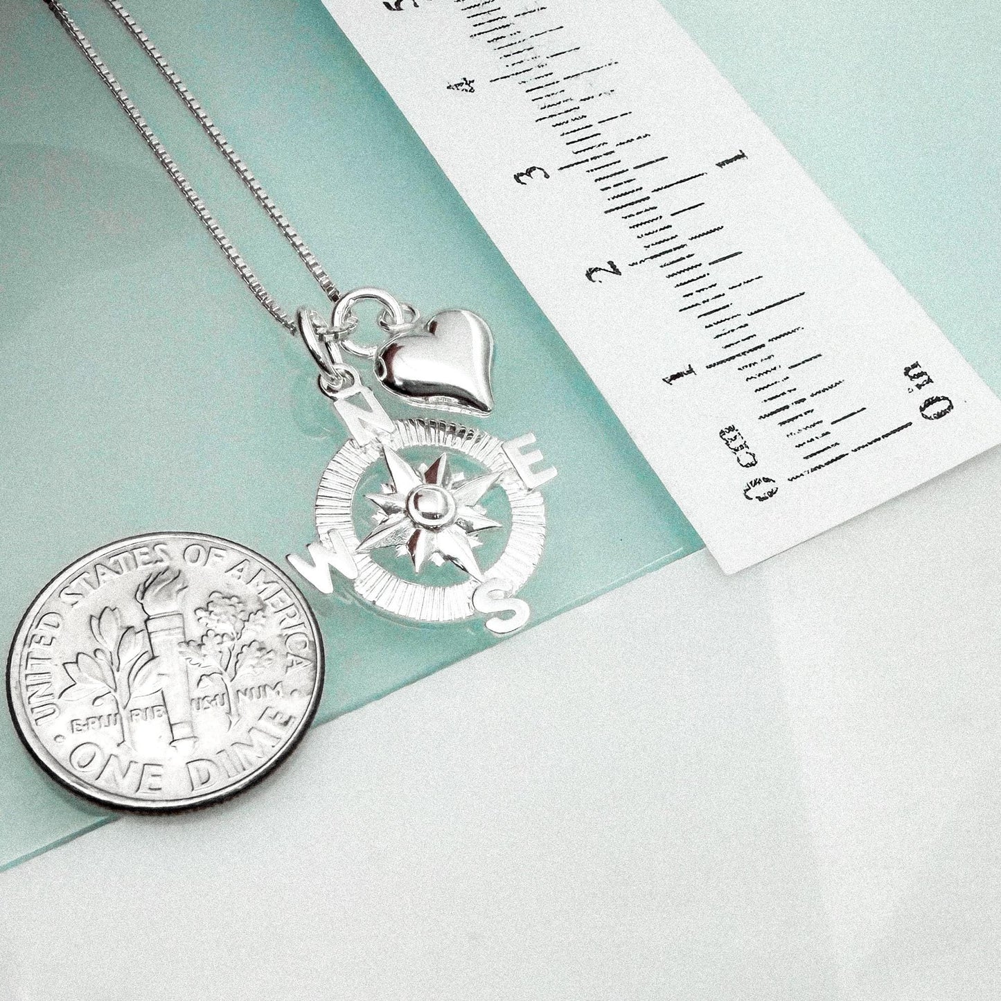 a silver compass pendant and a United States one-dollar coin, both placed on a light blue surface.