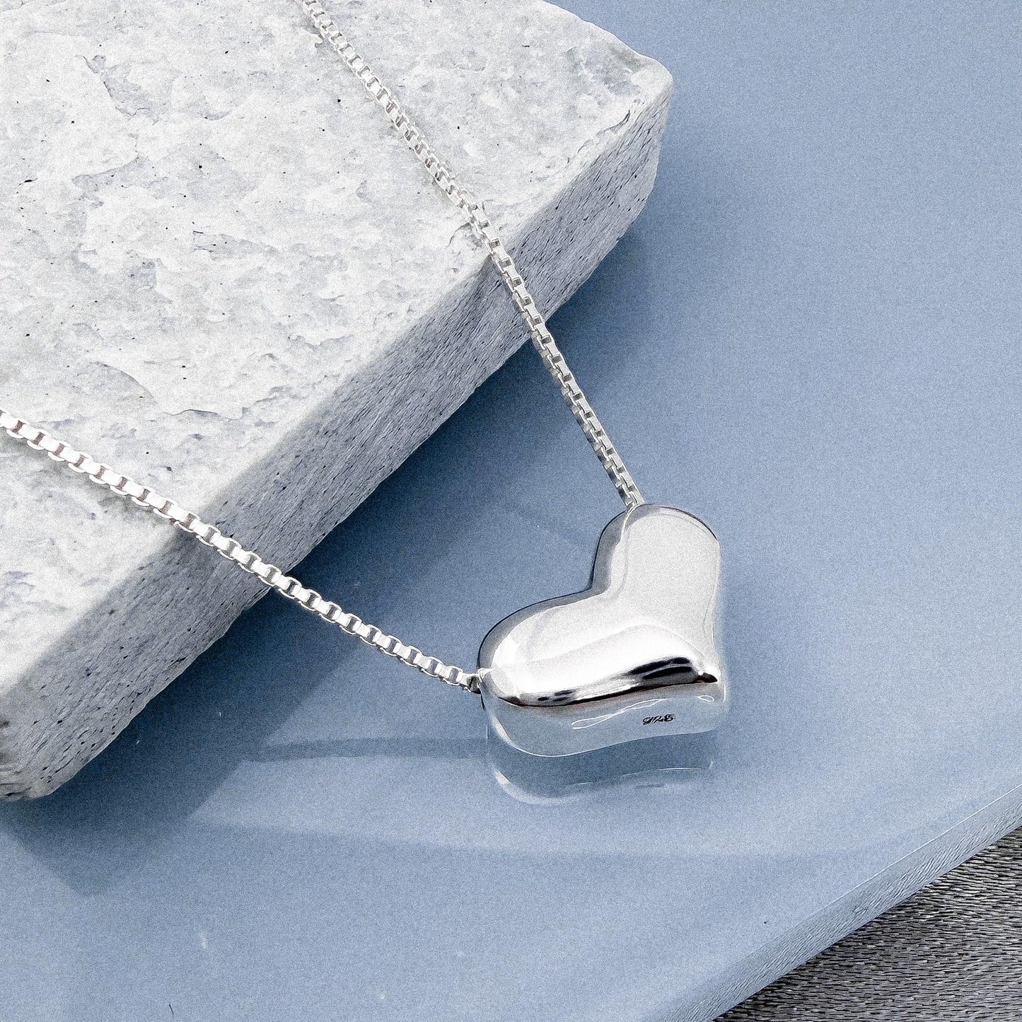 A silver heart-shaped pendant necklace is displayed on a gray stone surface.