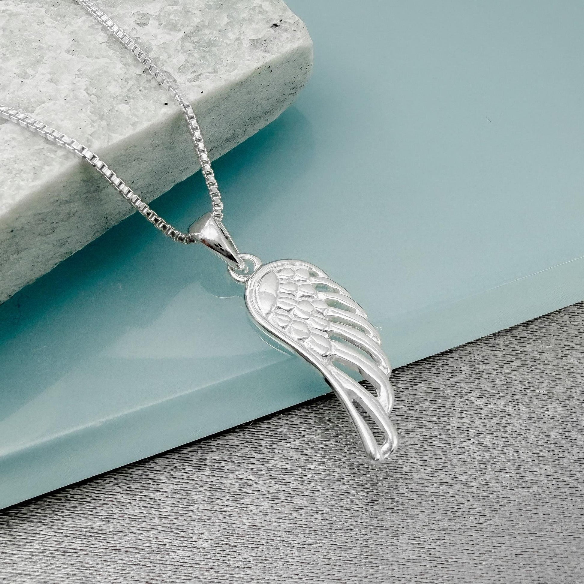 A silver-colored pendant necklace with a wing-shaped pendant is displayed on a gray surface.