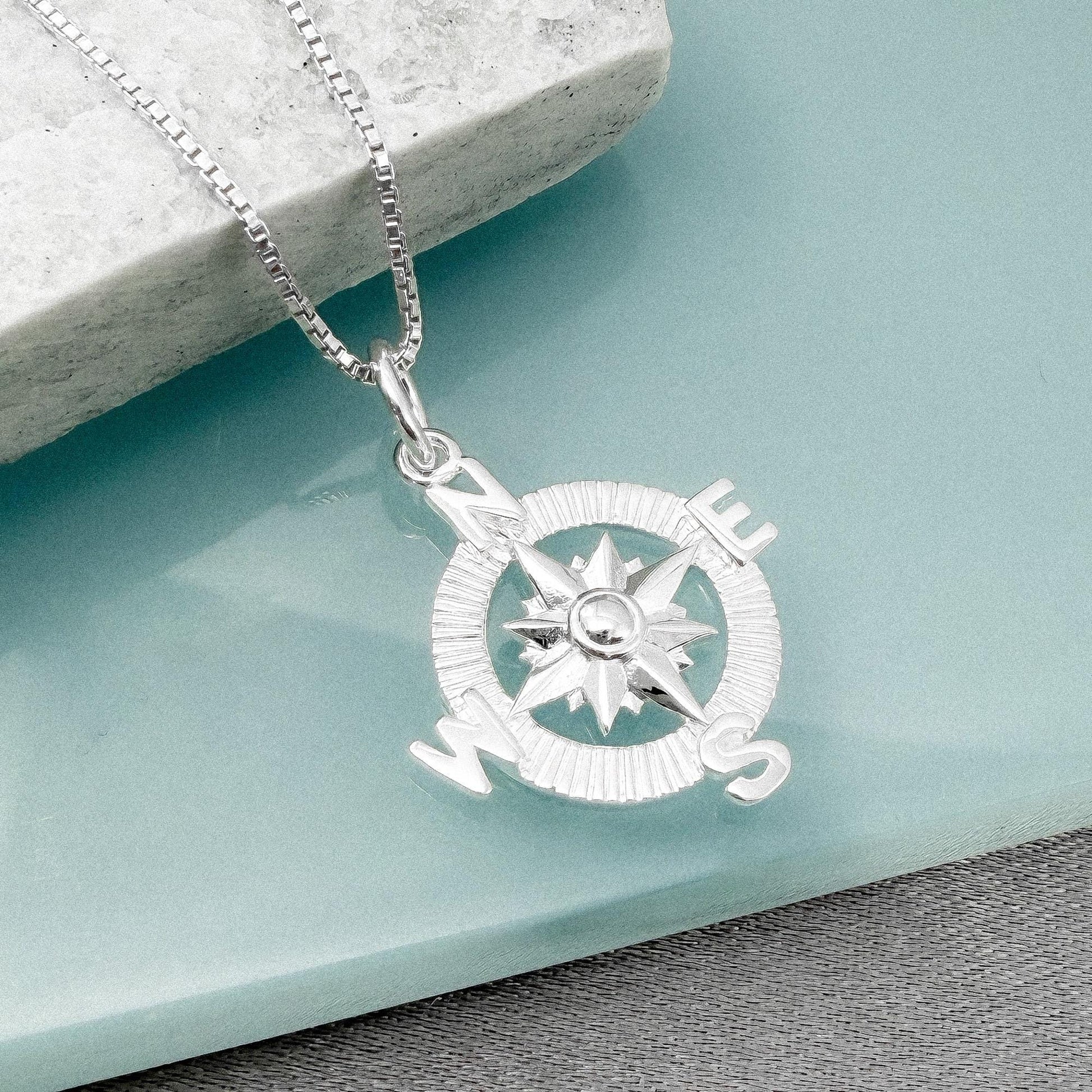A silver compass pendant with the letters "N", "E", "S", and "W" is displayed on a light blue surface.
