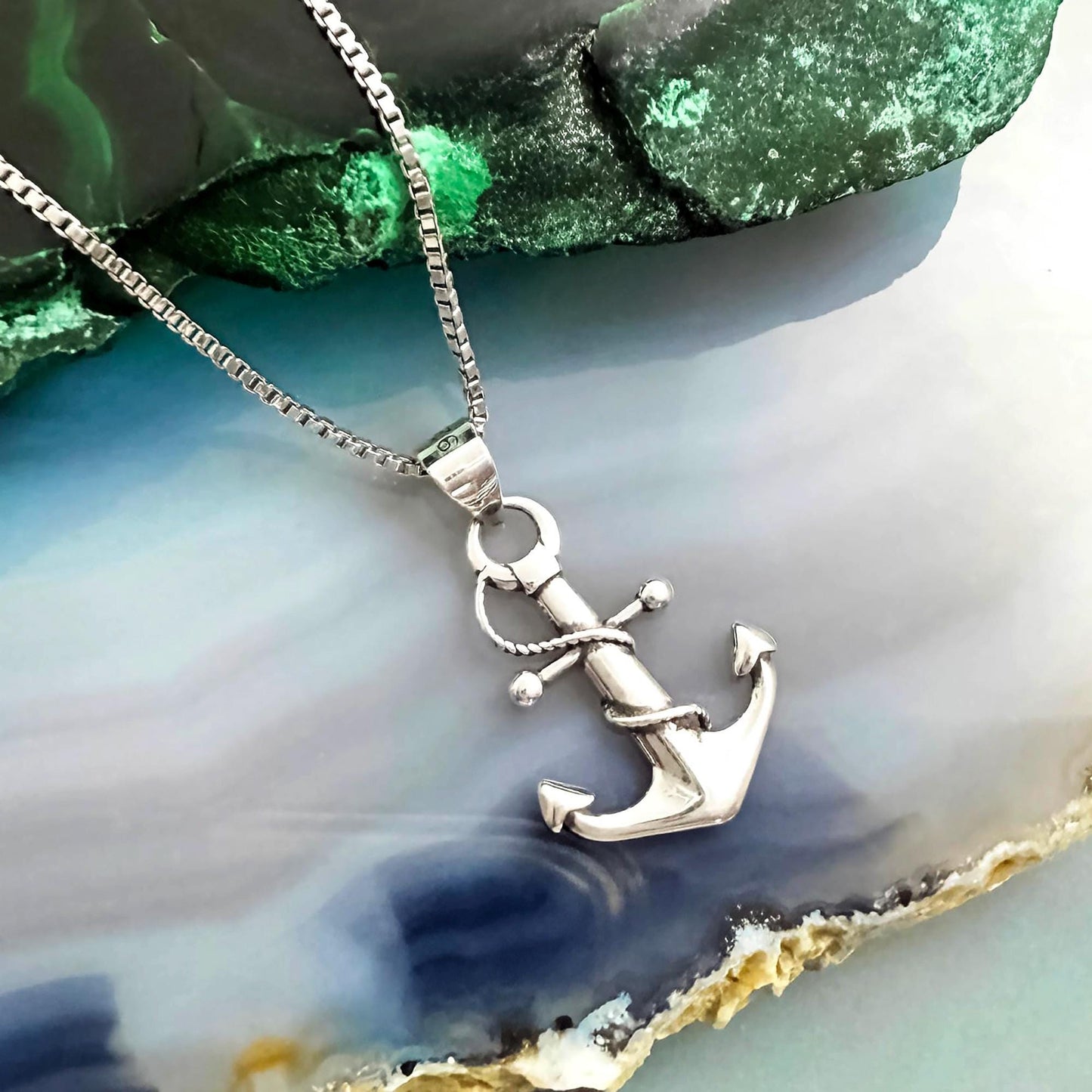 A silver anchor pendant necklace is displayed against a green and blue background.