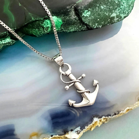 A silver anchor pendant necklace is displayed against a green and blue background.