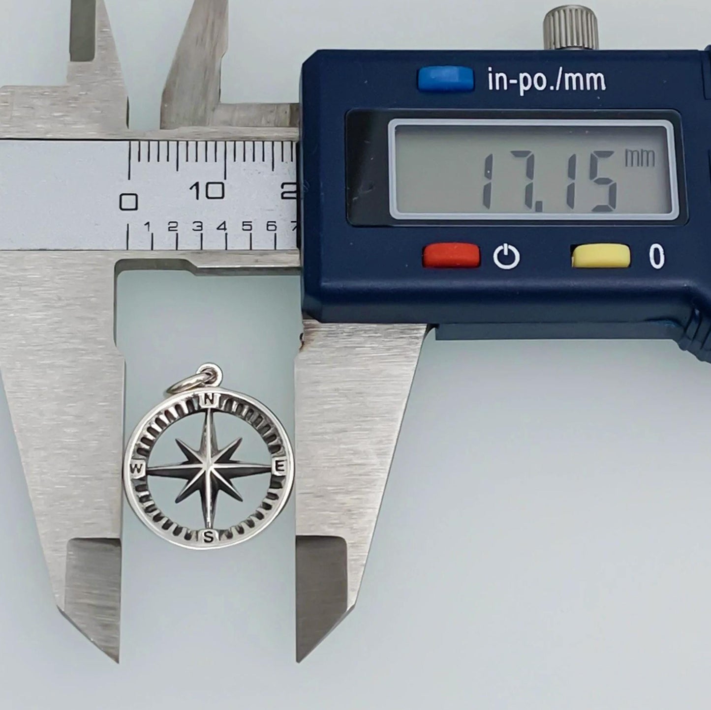 A silver compass charm is being measured with a caliper, which displays the size of the charm as 17.15 millimeters.