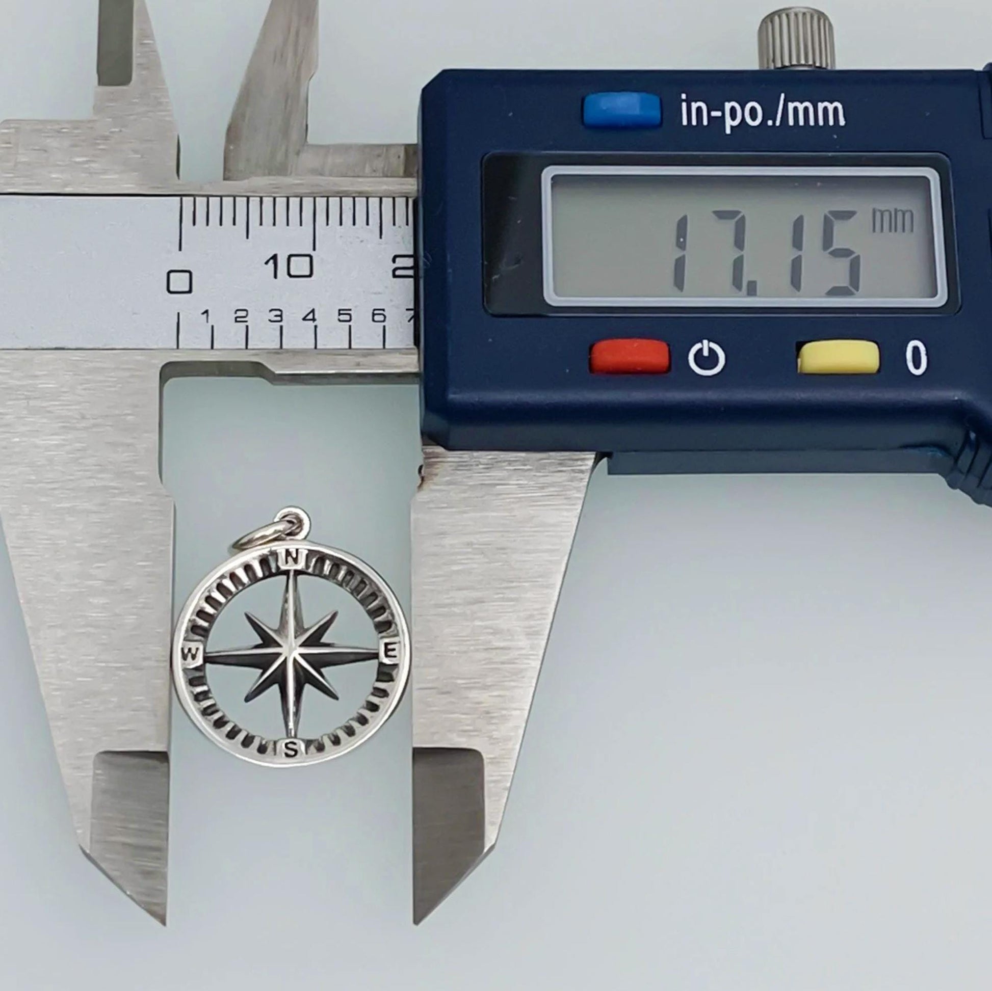 A silver compass charm is being measured with a caliper, which displays the size of the charm as 17.15 millimeters.