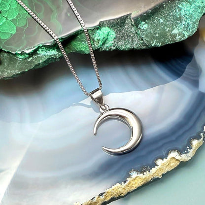 A silver crescent moon pendant necklace is displayed against a backdrop of a blue and green rock formation.