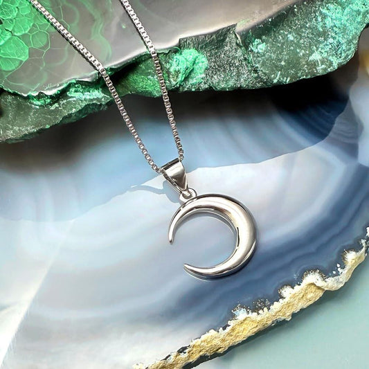 A silver crescent moon pendant necklace is displayed against a backdrop of a blue and green rock formation.