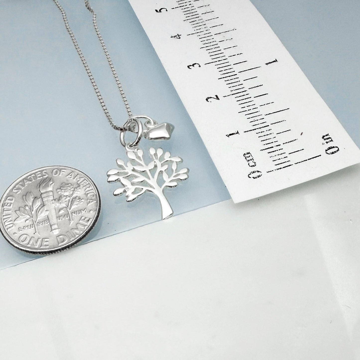 a silver tree pendant necklace and a U.S. quarter coin, with a ruler placed next to them.