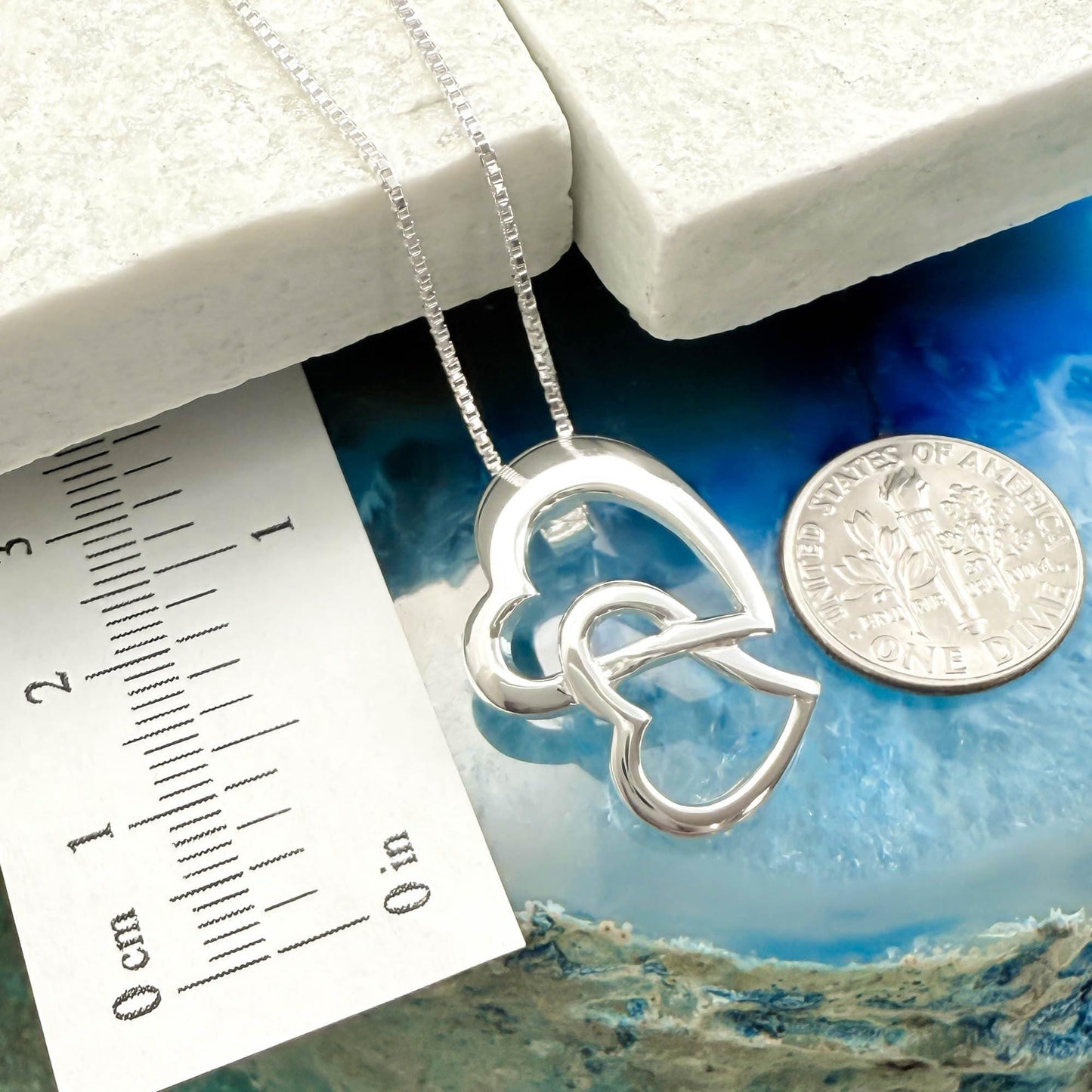a silver heart-shaped pendant necklace with a silver ruler placed next to it, and a United States quarter coin is also visible in the image.