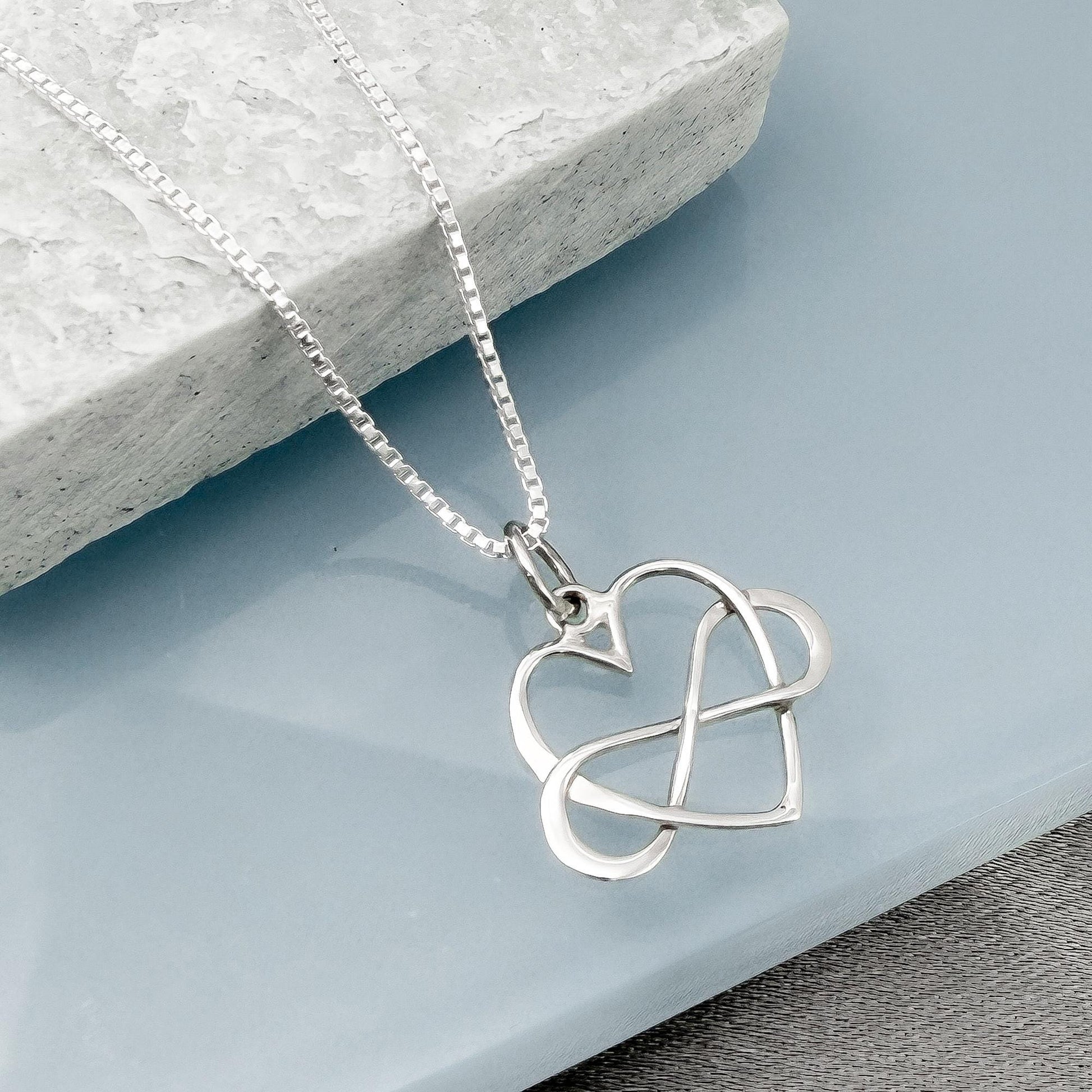 A silver heart-shaped pendant with a twisted knot design is displayed on a light blue surface.
