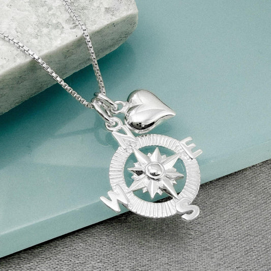 A silver compass pendant with a heart charm attached, resting on a light blue surface.