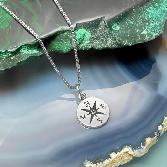A silver necklace with a compass pendant is displayed against a green and blue background.