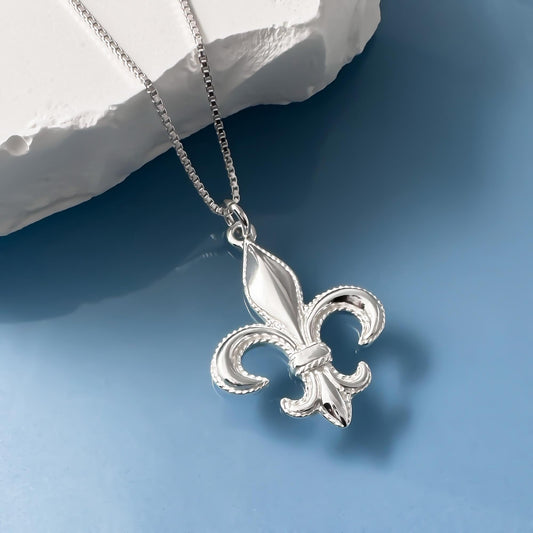 A silver-colored pendant in the shape of a fleur-de-lis, a heraldic symbol, is displayed on a chain against a blue background.