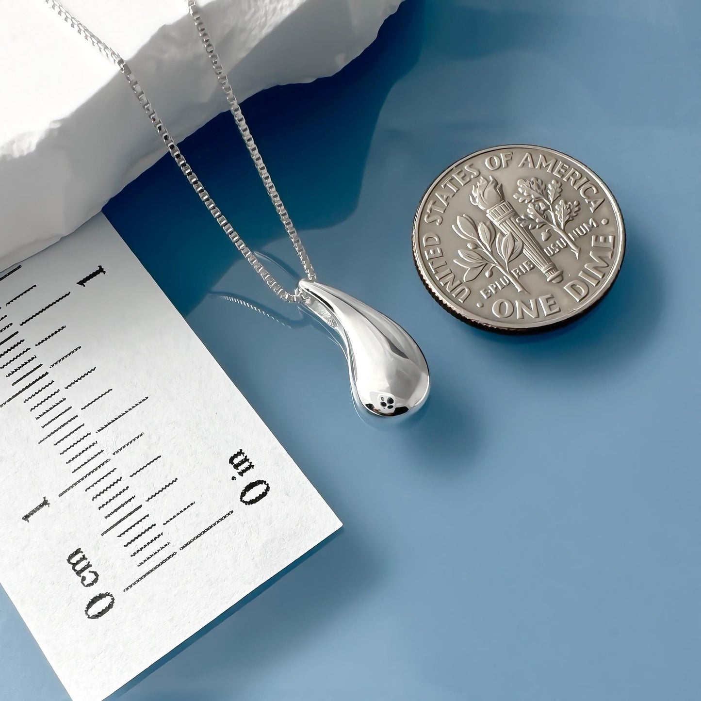 a silver necklace with a pendant, a U.S. one-dollar coin, and a paper with measurements on it, all placed on a blue surface.