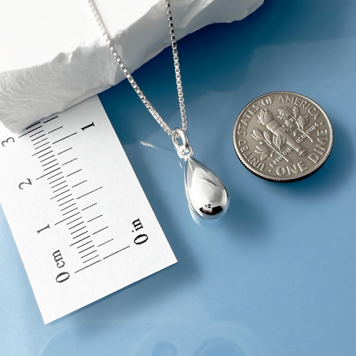 a silver necklace with a pendant, a silver coin, and a white paper with a scale for size comparison.