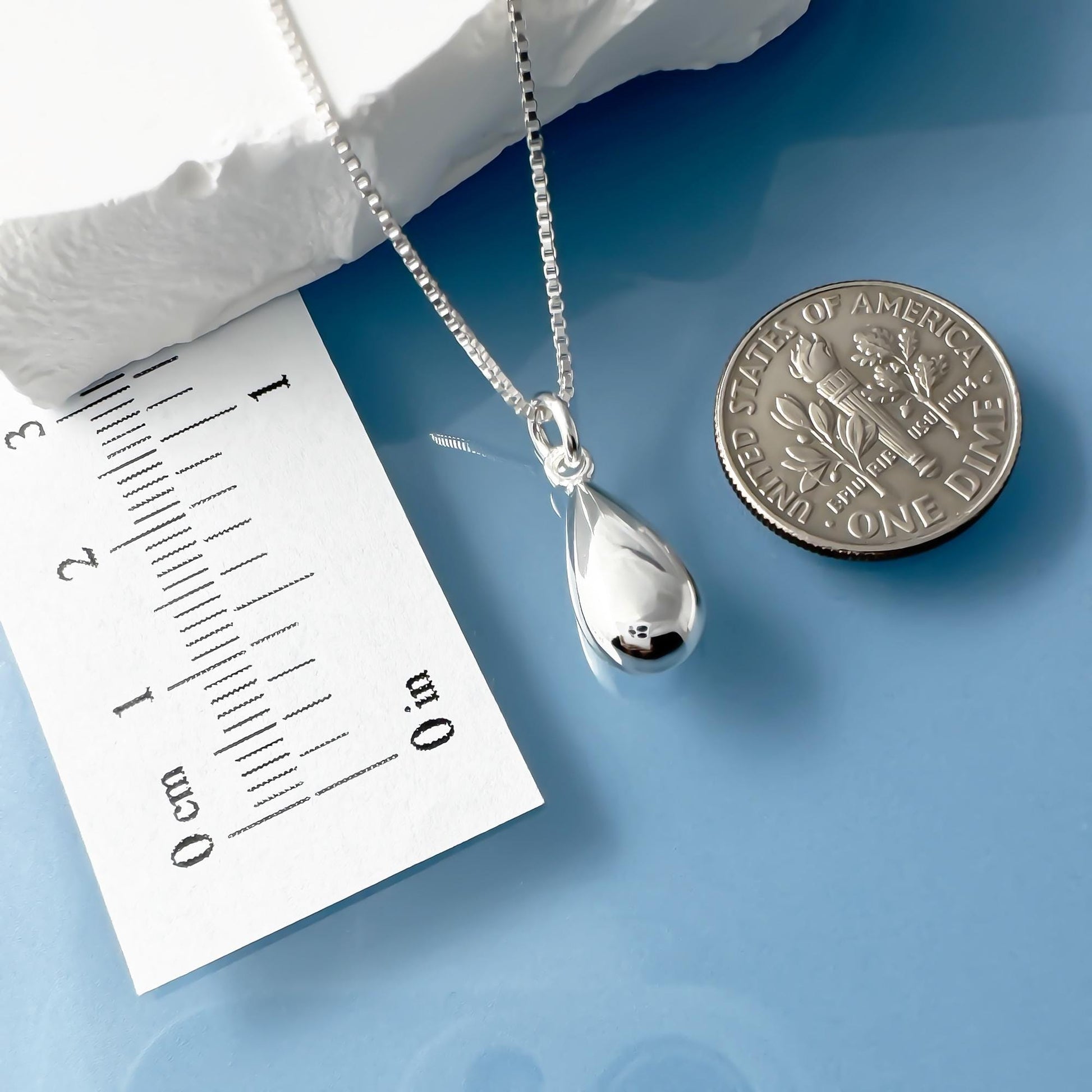 a silver necklace with a pendant, a silver coin, and a white paper with a scale for size comparison.