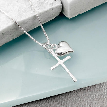 A silver cross pendant with a heart charm is displayed on a light blue surface.