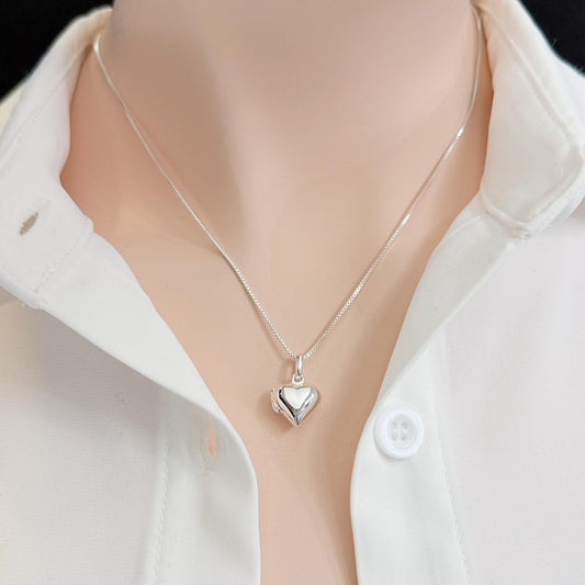 A silver heart-shaped pendant necklace is worn around a person's neck, with a white collared shirt visible underneath.