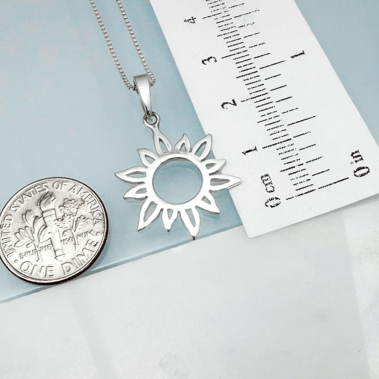 a silver pendant necklace with a sun-shaped design, placed next to a United States quarter coin and a ruler for size comparison.