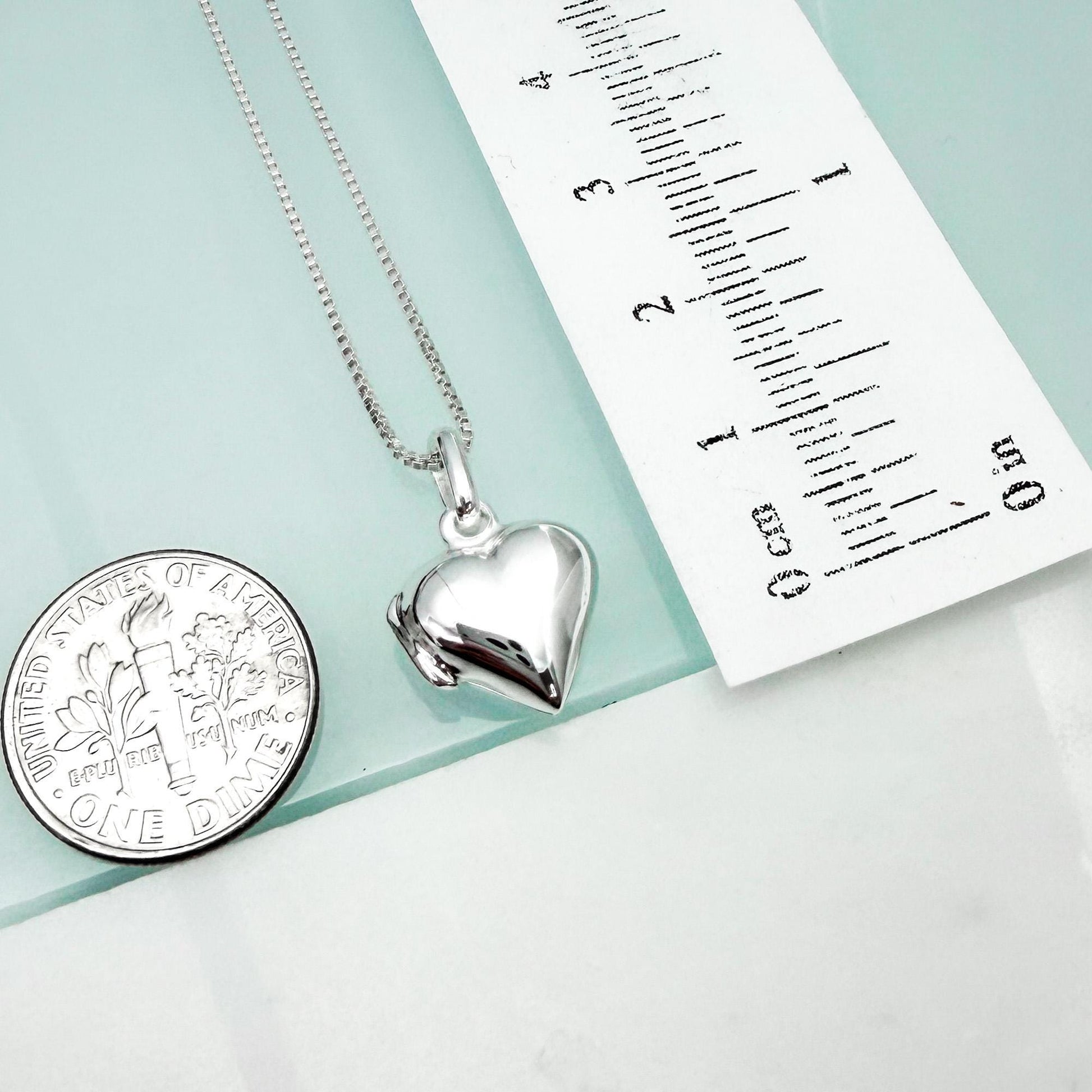 a silver heart-shaped locket pendant and a United States quarter coin, both placed on a light blue surface next to a ruler for size comparison.