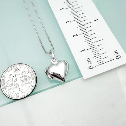 a silver heart-shaped locket pendant and a United States quarter coin, both placed on a light blue surface next to a ruler for size comparison.