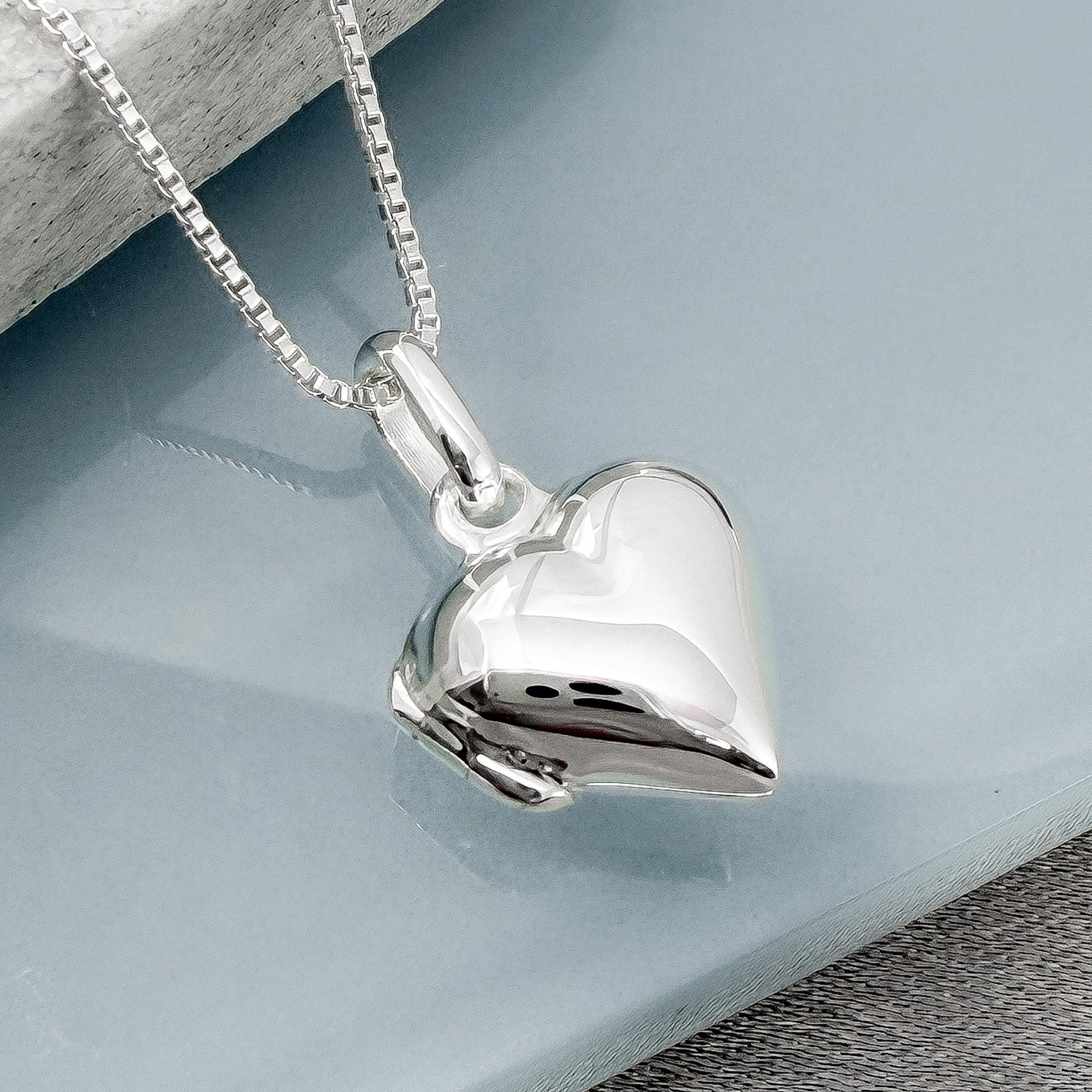 A silver heart-shaped pendant necklace with a chain is displayed on a blue surface.