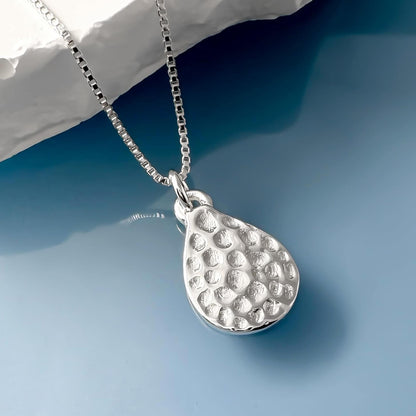 A silver necklace with a pendant in the shape of a teardrop is displayed against a blue background.
