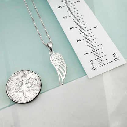 a silver pendant with a wing design, a silver coin, and a ruler on a light blue background.