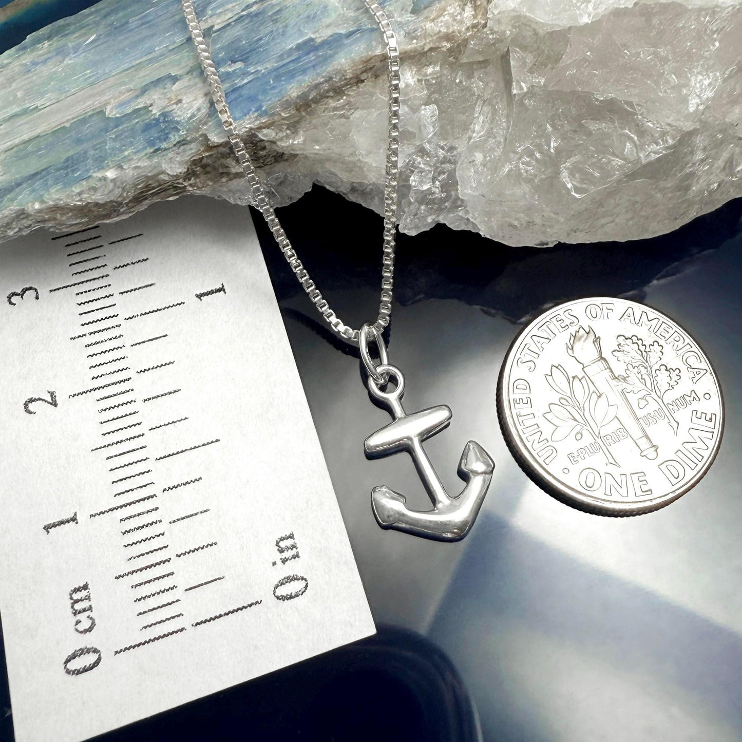 a silver anchor pendant hanging from a chain, placed next to a United States one dollar coin.