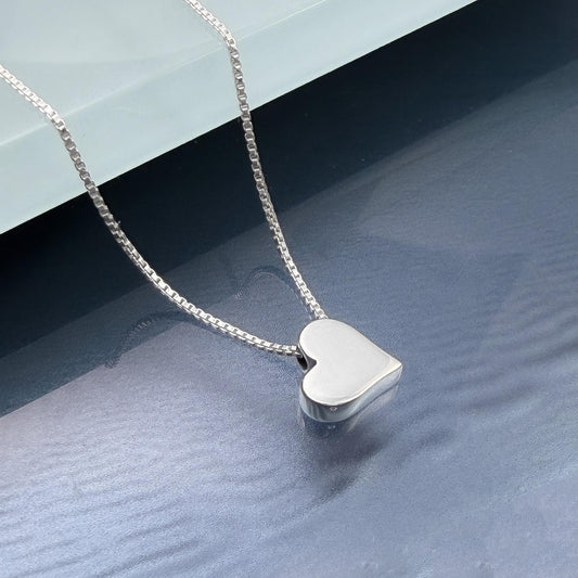 A silver heart-shaped pendant necklace is displayed on a reflective surface.
