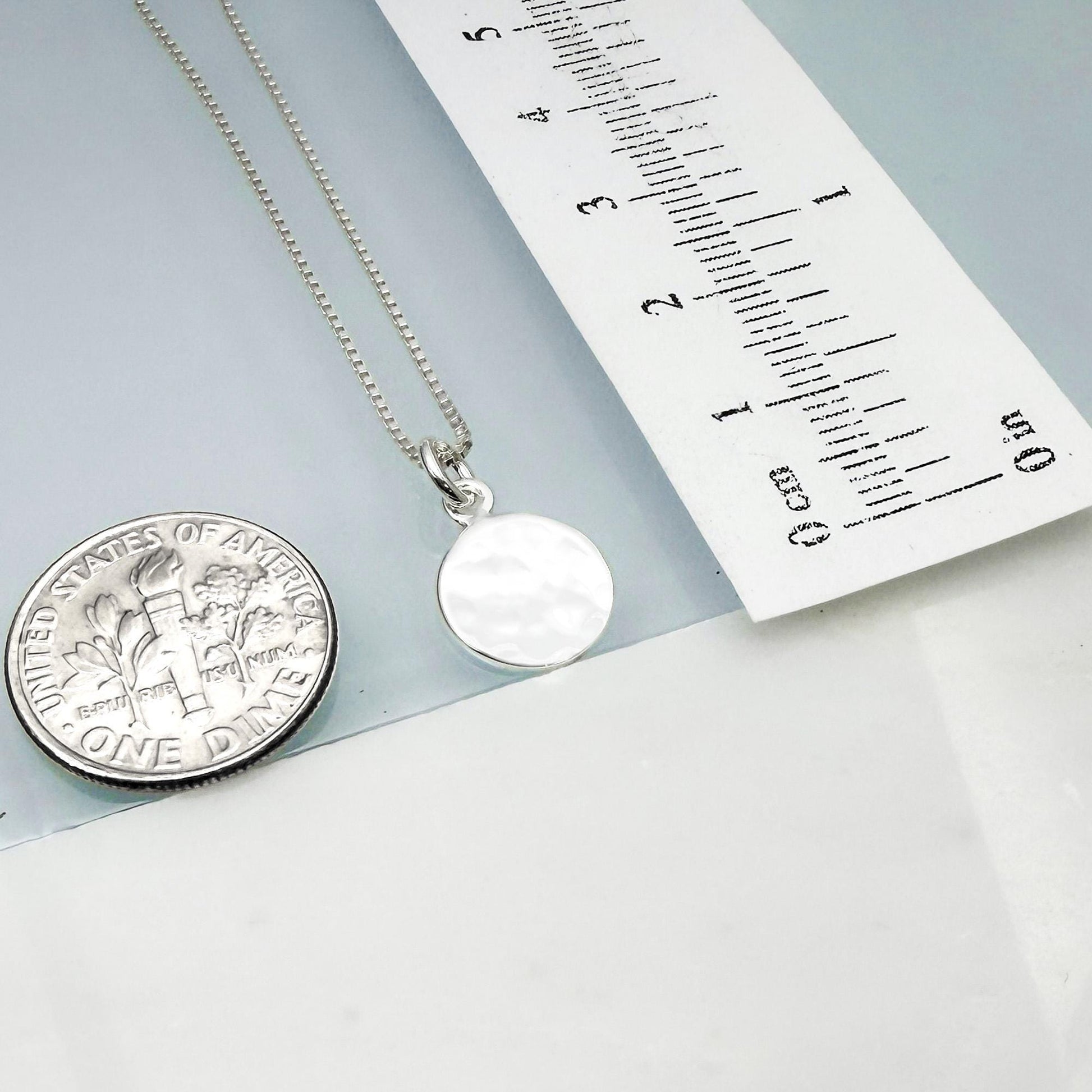 a silver necklace with a round pendant, placed next to a United States one dollar coin for size comparison.