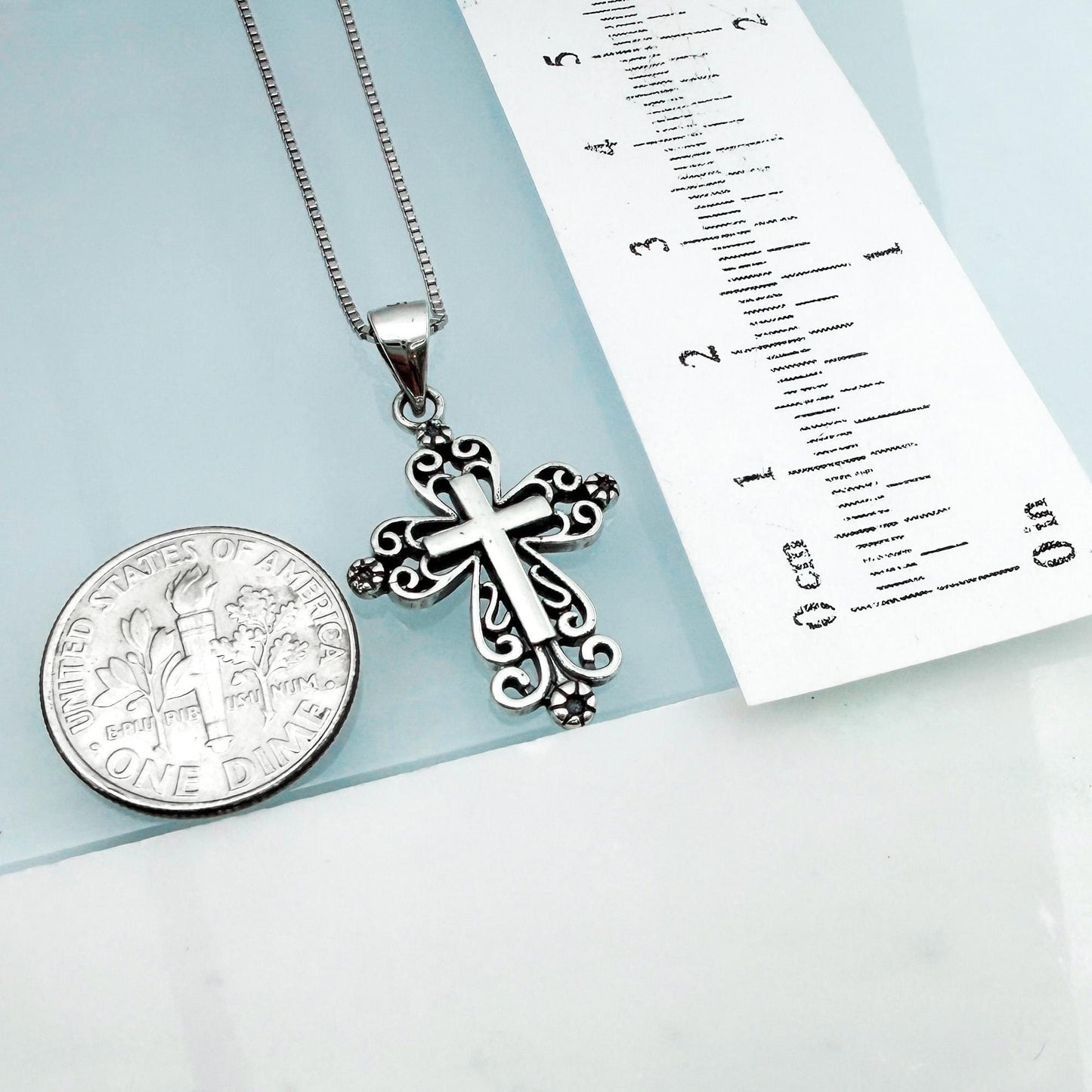 a silver cross pendant hanging from a chain, placed next to a United States one-dollar coin.
