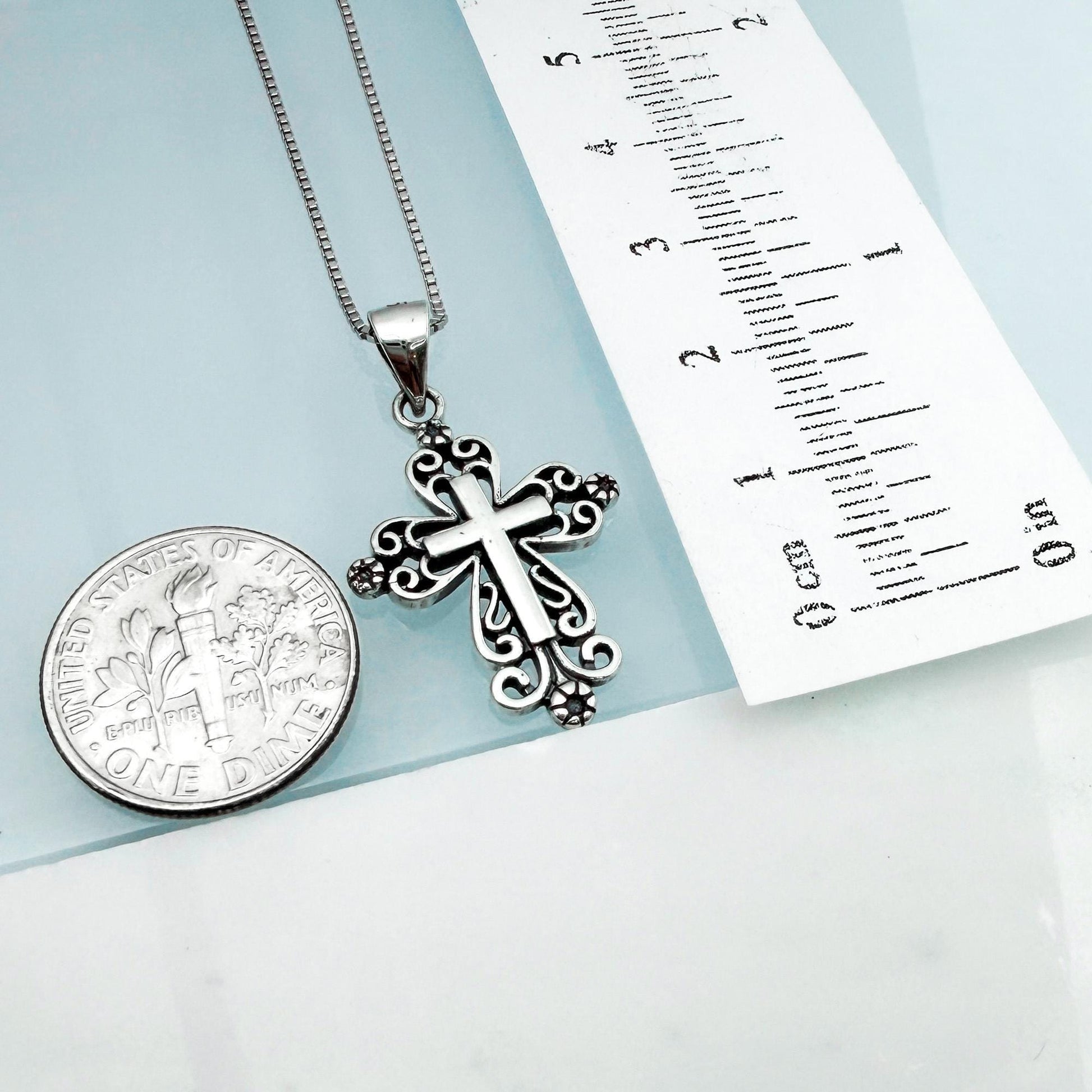 a silver cross pendant hanging from a chain, placed next to a United States one-dollar coin.