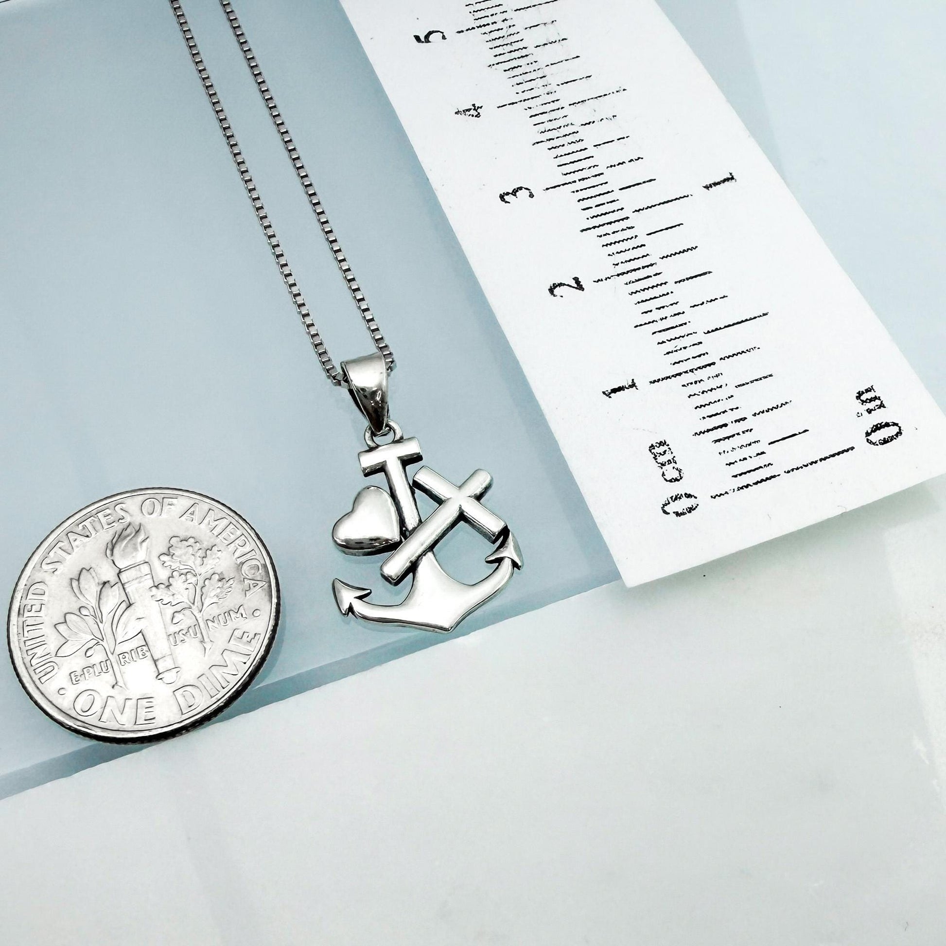 a silver necklace with a heart-shaped pendant and an anchor charm, placed next to a United States one-dollar coin.